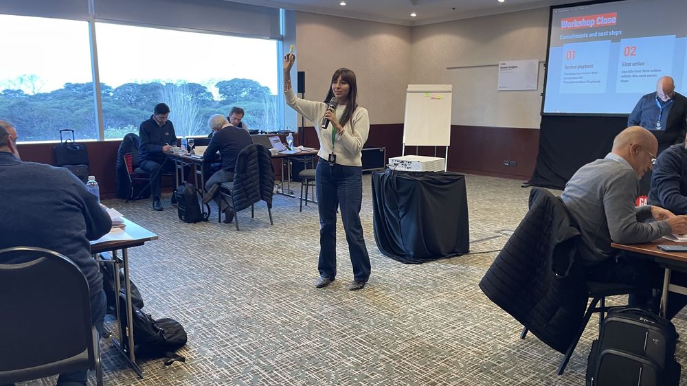 Alejandra Endara facilitating a corporate AI workshop, one hand raised, with participants working at tables